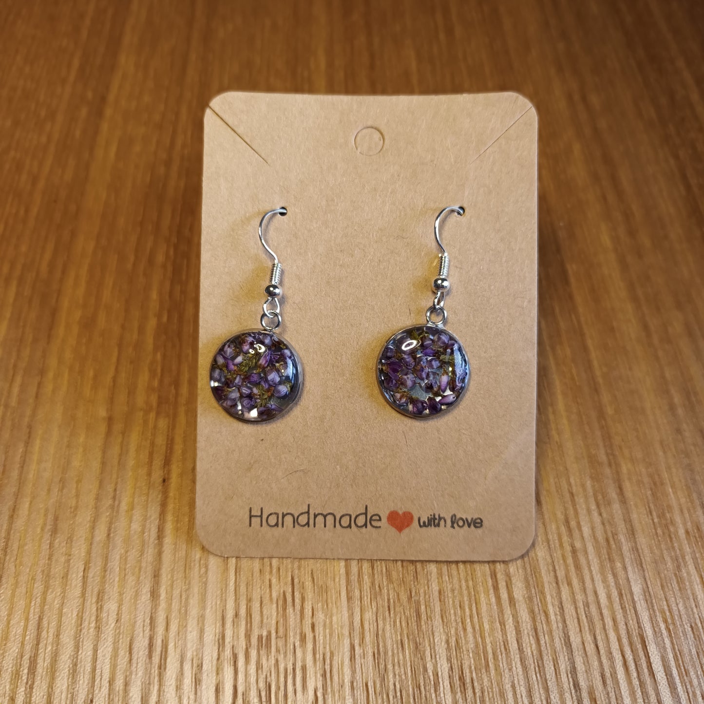 Pair of purple floral earrings on a brown card with 'Handmade with Love' text, placed on a wooden surface.